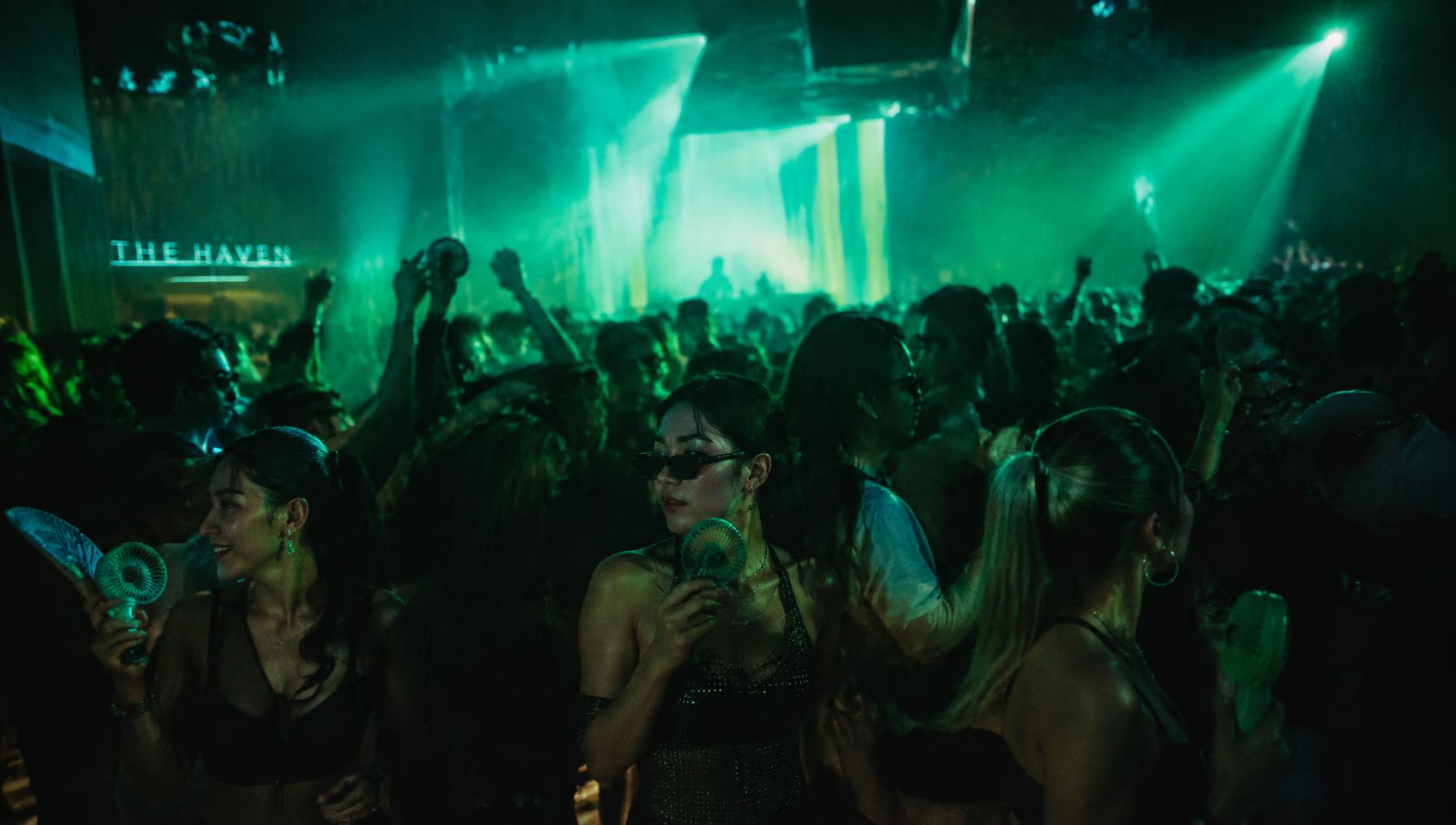 A packed nightclub dance floor under green lights, where guests cool off with handheld fans.